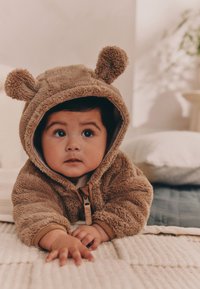 Sweat à capuche zippé en polaire marron avec des oreilles d'ours, présentant une texture douce et des poignets côtelés. L'enfant est allongé sur une surface texturée.