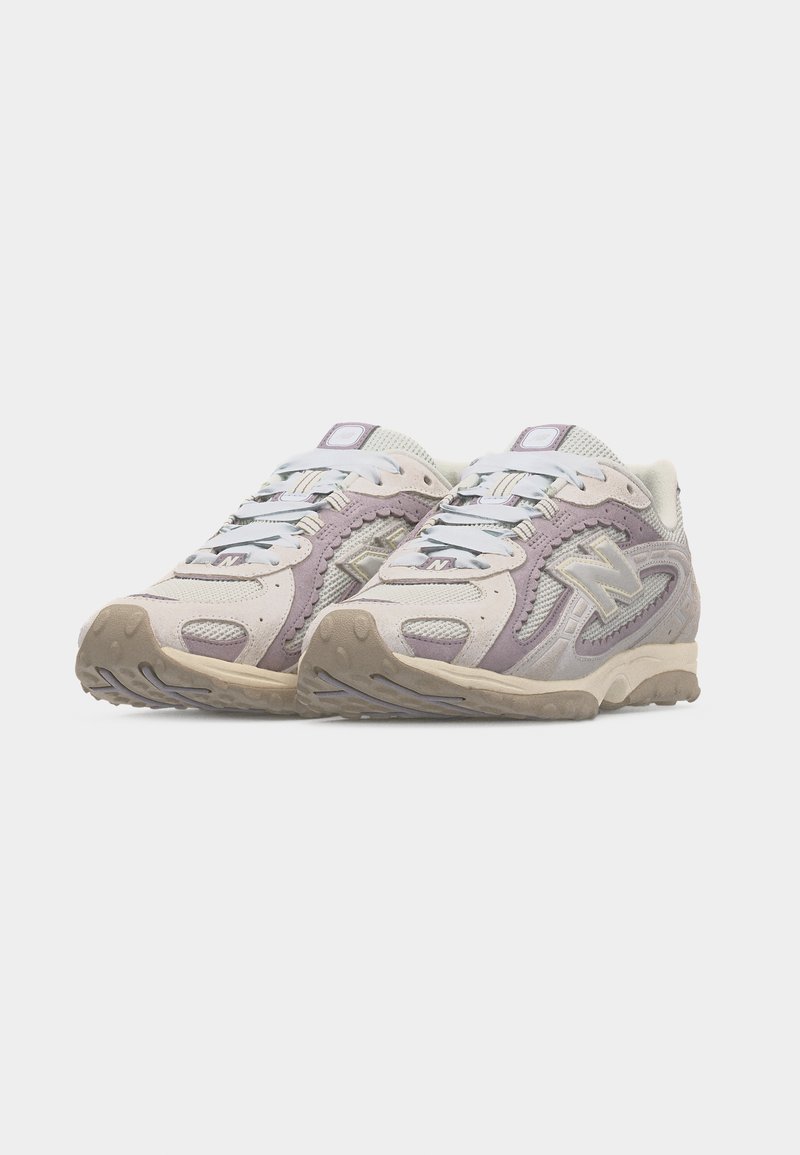 Par de zapatillas New Balance en beige y violeta claro con detalles en malla y ante, cordones blancos y suelas de goma beige sobre un fondo blanco.