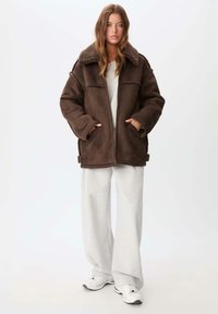 Giovane donna in piedi con indosso un giubbotto oversize in shearling marrone, pantaloni felpati bianchi larghi e sneakers bianche contro uno sfondo bianco.