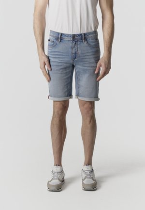 Mann trägt hellblau hochgekrempelte Jeansshorts, weißes T-Shirt und weiße Sneaker, steht vor einem schlichten hellen Hintergrund.