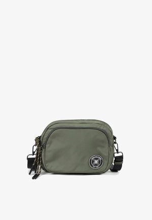 Bolso bandolera compacto de color verde oliva con dos compartimentos con cremallera, correa negra ajustable y parche redondo con logo en blanco y negro.