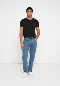 Camiseta negra de manga corta, jeans de mezclilla azul, zapatillas blancas y cinturón negro; modelo posa con las manos en los bolsillos, de pie contra un fondo neutro.