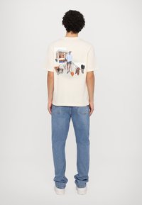 Kremfarget bomulls t-skjorte med et fargerikt grafisk motiv av en person som går flere hunder mot bakgrunn av en dyrebutikk. Løs passform og korte ermer.