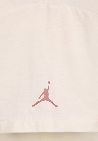 Camiseta de algodón crema con un logo Jumpman rosa en el dobladillo. Textura suave con un diseño moderno y minimalista.