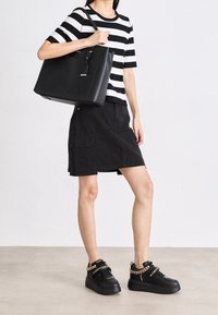 Personne portant une chemise rayée noire et blanche, une jupe noire, des baskets plateformes noires avec des chaînes dorées, tenant un grand sac fourre-tout noir sur l'épaule.