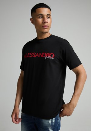 Ung mand iført en sort t-shirt med rød tekst "ALESSANDRO Milano" og blå slidte jeans, stående mod en ensfarvet lys baggrund.