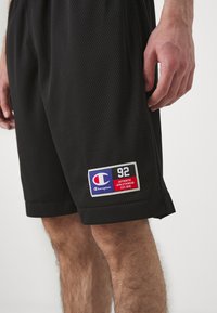 Pantalones cortos deportivos de malla negra con bolsillos laterales, cinturilla elástica y un parche de logo de Champion con acentos rojos y azules.