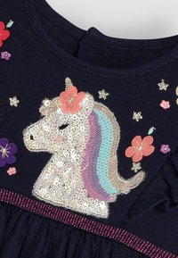 Kleid in Marineblau mit einem Pailletten-Einhorn-Design, buntem Mähnen, floralen Applikationen und einem lila Akzentband in der Taille.