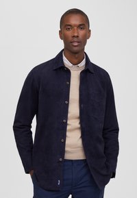 Chaqueta de ante azul marino con cierre de botones, ligero brillo, cuello y bolsillo en el pecho. Superpuesta sobre un suéter beige, combinada con pantalones de cuadros.