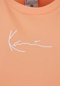 Peachfarget T-skjorte med ribbet tekstur og rund hals, med et hvitt brodert signaturdesign på forsiden.