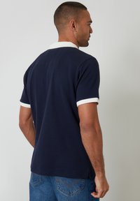 Hombre de perfil, vistiendo polo azul marino de manga corta con cuello y puños blancos, y vaqueros azules sobre un fondo claro liso.