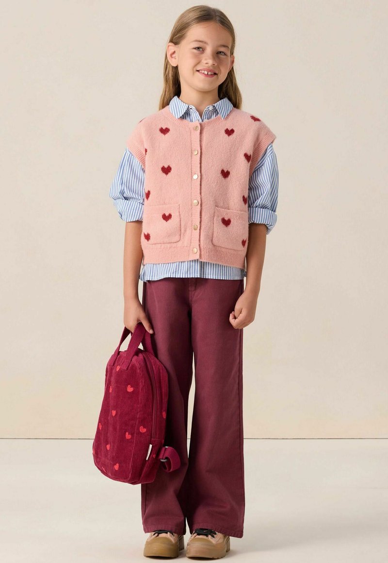 Une jeune fille portant un gilet sans manches rose avec des cœurs rouges, un t-shirt rayé, un pantalon bordeaux, tenant un sac à dos assorti à motif de cœurs.