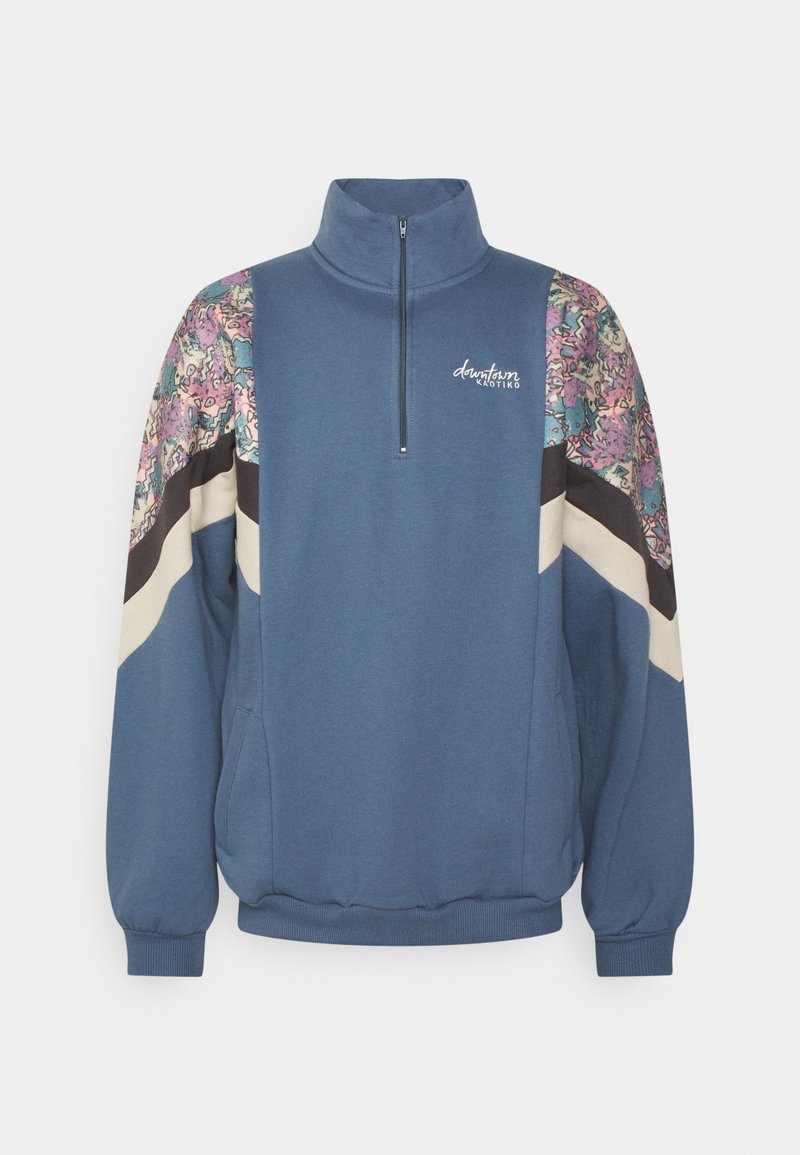 Pull bleu à manches longues avec demi-fermeture éclair, panneaux d'épaules à motifs aux couleurs pastel, rayures chevron noires et blanches, et texte « downtown KAOTIKO » sur la poitrine.
