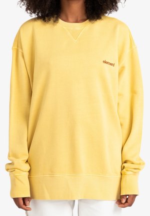 Person iført en løstsiddende gul sweatshirt med lille brunt "element"-logo på venstre bryst, kombineret med hvide bukser.