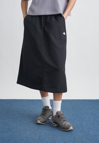 Άτομο που φοράει μαύρη φούστα μέσου μήκους, γκρι αθλητικά παπούτσια, λευκές κάλτσες και γκρι μπλούζα, με τα χέρια στις τσέπες, όρθιο πάνω σε μπλε χαλί.