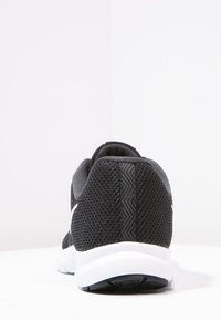 Černá atletická obuv z síťoviny z pohybu zezadu, s bílou podrážkou a jemným texturovaným vzorem na patě na bílém pozadí.