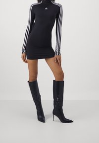 Czarna sukienka z długim rękawem, z kołnierzykiem typu mock oraz białymi paskami; w połączeniu z czarnymi, wysokimi na kolano botkami na szpic z zamkiem.