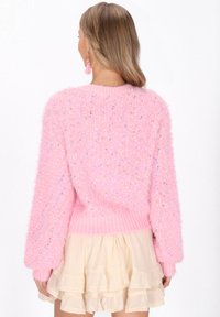 Egy világosbarna hajú nő, aki pihe-puha rózsaszín pulcsit és tejszínszínű, réteges mini szoknyát visel, hátulról látható.