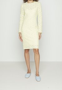 Kvinna som bär en knälång, långärmad klänning i ljusgul spets, kombinerad med ljusblå, vävda platåskor, stående mot en enkel vit bakgrund.