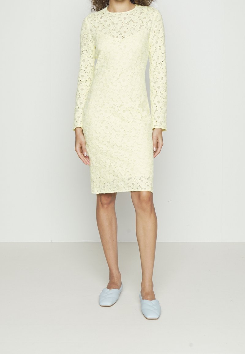 Kvinna som bär en knälång, långärmad klänning i ljusgul spets, kombinerad med ljusblå, vävda platåskor, stående mot en enkel vit bakgrund.