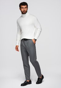 Biały sweter z wysokim kołnierzem, szare dopasowane spodnie i czarne buty eleganckie. Zauważalny zegarek na nadgarstku, w pozycji stojącej na neutralnym tle.