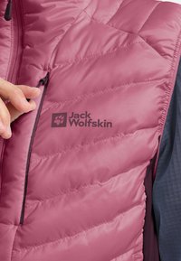 Gilet rosa trapuntato con tasca con zip, con il logo Jack Wolfskin. Il materiale appare liscio con una finitura leggermente lucida.