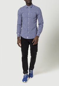 Mans långärmad skjorta i marinblå och vit rutigt mönster, knapp-up design, kombinerad med svarta jeans och blå sneakers.