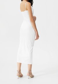 Bílá, přiléhavá midi šaty s tenkými ramínky, hladkém povrchu a bočním řasením. Doplněno stříbrnými sandály na vysokém podpatku s úzkým podpatkem.