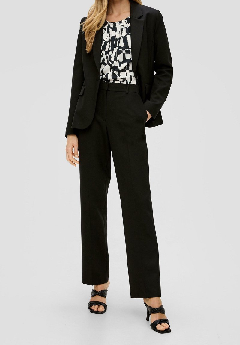 Black structured blazer over a patterned white and black blouse, paired with black straight-leg trousers and black strappy heels.