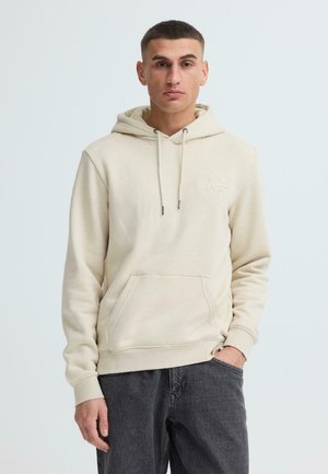 Jeune homme aux cheveux courts portant un sweat à capuche beige avec poche avant et un jean gris foncé, debout devant un fond clair uni.