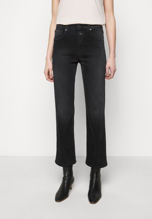 Vrouw die zwarte, hooggesloten jeans met rechte pijpen draagt, gecombineerd met zwarte enkellaarsjes met hak, staand tegen een effen lichte achtergrond.