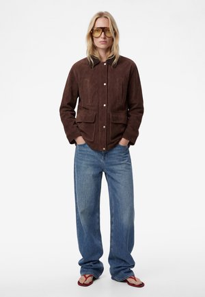 Frau trägt übergroße braune Wildlederjacke, weite blaue Jeans, bernsteinfarbene Sonnenbrille und rote Flip-Flops, steht mit Händen in den Taschen.