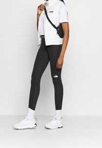 Des leggings noirs avec un logo blanc, associés à un gilet rembourré blanc et un t-shirt blanc simple. Des chaussures de sport aux teintes neutres complètent le look.