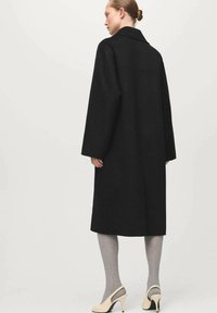 Czarny wełniany płaszcz o długiej, prostej sylwetce, szerokich rękawach i dużym kołnierzu. Modelka ma na sobie szare rajstopy i beżowe buty na wysokim obcasie.