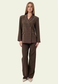 Mujer de pie con blazer marrón de doble botonadura y pantalones anchos a juego, con zapatos de tacón beige puntiagudos sobre fondo claro.