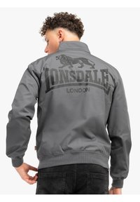 Siva bomber jakna s mat finishem, koja ima veliki crni logo lava i tekst "LONSDALE LONDON" na leđima, rebrasti rukavi i donji deo.
