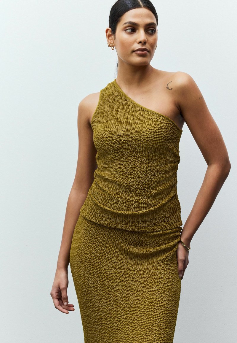 Vestido de punto de un solo hombro en verde oliva, con una tela texturizada y detalles fruncidos en la cintura y el dobladillo. Diseño y ajuste simples.