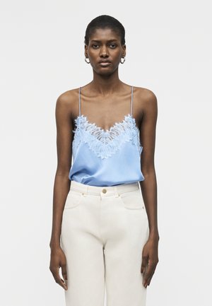 Moteris, vilkinti šviesiai mėlyną nėriniais puoštą palaidinę su plonomis petnešėlėmis ir baltais aukštakulniais kelnėmis, stovinti priešais paprastą šviesų foną.