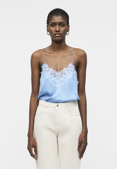 Femme portant un caraco bleu clair orné de dentelle avec de fines bretelles et un pantalon blanc taille haute, debout devant un fond clair uni.