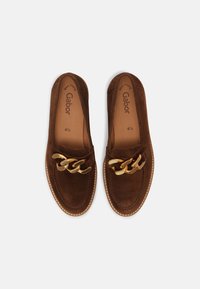 Zapatos de ante marrón tipo slip-on con un acento de cadena dorada texturizada en el empeine y costuras blancas contrastantes alrededor de la suela.