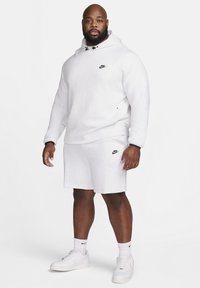Balts Nike kapucis krekls ar melnu logo, saskaņots ar pelēkiem šortiem un baltām sporta kurpēm. Ikdienišķs izskats ar kapuci un stinguma auklām.