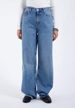 Jean large en denim bleu clair, taille haute avec des coutures visibles, poches avant et fermeture à bouton. Porté avec des chaussures noires.