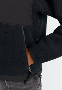 Hand i sidoficka på svart fleecetröja med dragkedja, texturerat tyg och justerbara fållstopp, bärs med grå denimjeans.