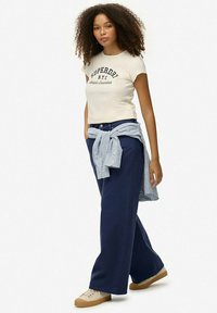 Camiseta ajustada beige con el texto "SUPERDRY". Pantalones anchos de color azul marino. Camisa a rayas azul clara atada a la cintura. Zapatillas color beige con suela texturizada.