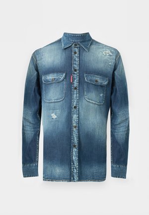 Homme portant une chemise en denim délavée sur un débardeur blanc, un short cargo en denim bleu clair, des chaussettes blanches et des baskets vert foncé, debout avec la main dans la poche.