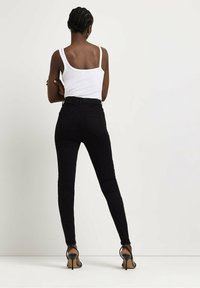 Jeans ajustados de tiro alto negros con una textura suave, combinados con una camiseta sin mangas blanca de canalé. La modelo lleva tacones negros con tiras, vista desde atrás.