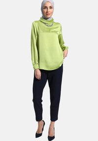 Grüne Satinbluse mit hohem Kragen, langen Ärmeln und Knopfmanschetten; kombiniert mit einer navyblauen, taillierten Hose und navyblauen Absätzen. Silberne Halskette als Akzent.