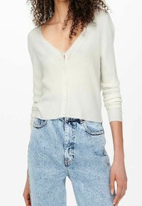 Cienki, prążkowany, biały kardigan zapinany na guziki z dekoltem w serek noszony z jasnoniebieskimi, wysokiej talii dżinsami z efektem sprania przez osobę z kręconymi włosami.