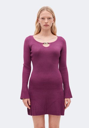 Femme portant une robe violette ajustée côtelée à manches longues avec des poignets évasés et un petit fermoir métallique au niveau de l'encolure.