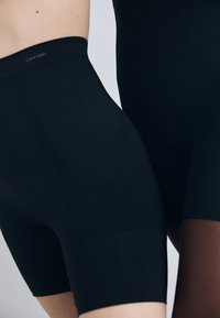 Crne tajice od rastezljivog materijala, s glatkom teksturom, širokim pojasom s logom "Calvin Klein", duljine do sredine bedara i minimalističkog dizajna.
